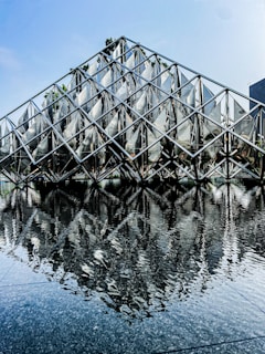 A geometric structure with a triangular metal framework stands in front of a reflective water surface, creating a symmetrical reflection. The structure's metallic elements are arranged in a diamond pattern, and small trees are visible in the background. The sky is clear with a light blue hue.