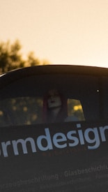 A person with red hair is partially visible through the rear window of a vehicle. The focus is on the text 'design' on the vehicle, which is part of a larger sign or advertisement. The lighting suggests a sunset or sunrise, with a warm, golden hue dominating the background.