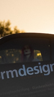 A person with red hair is partially visible through the rear window of a vehicle. The focus is on the text 'design' on the vehicle, which is part of a larger sign or advertisement. The lighting suggests a sunset or sunrise, with a warm, golden hue dominating the background.