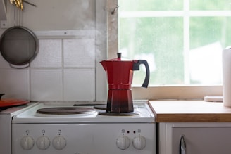 A red stovetop espresso maker is placed on a white electric stove in a kitchen. Steam is rising from the pot. To the left, a wall-mounted frying pan can be seen. A roll of paper towels is on the countertop next to the stove, and sunlight filters through a window with a view of greenery.