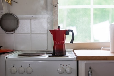 A red stovetop espresso maker is placed on a white electric stove in a kitchen. Steam is rising from the pot. To the left, a wall-mounted frying pan can be seen. A roll of paper towels is on the countertop next to the stove, and sunlight filters through a window with a view of greenery.