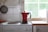 A red stovetop espresso maker is placed on a white electric stove in a kitchen. Steam is rising from the pot. To the left, a wall-mounted frying pan can be seen. A roll of paper towels is on the countertop next to the stove, and sunlight filters through a window with a view of greenery.