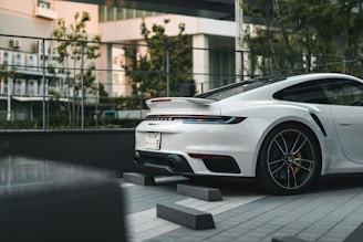 A white sports car is parked on a tiled surface in an outdoor urban setting. The car has a sleek design with a visible rear spoiler and dark-tinted windows. In the background, there's a modern building with large windows and some trees and plants. Barriers are placed around the vehicle.
