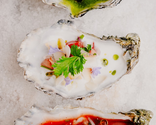 A close-up of fresh oysters resting on a bed of coarse salt. The oyster at the center is garnished with vibrant toppings, including parsley, green herbs, a slice of tomato, and delicate purple flowers. The natural textures of the oyster’s shell contrast with the smoothness of the toppings.