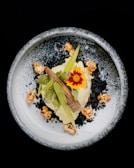 An artistic food presentation featuring a white plate with a decorative border, topped with a gourmet dish. The central focus is a vibrant red and yellow flower, surrounded by leafy greens, popcorn, and a piece resembling a small cob of corn. Black and white crumb-like elements enhance the visual appeal.
