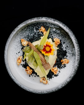An artistic food presentation featuring a white plate with a decorative border, topped with a gourmet dish. The central focus is a vibrant red and yellow flower, surrounded by leafy greens, popcorn, and a piece resembling a small cob of corn. Black and white crumb-like elements enhance the visual appeal.