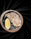 A single oyster on a half shell rests on a bed of ice inside a stone bowl, accompanied by a slice of lemon. The backdrop is a smooth, dark fabric that adds a touch of elegance to the composition.