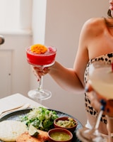 A person holding a vibrant red cocktail garnished with an orange flower, with a plate of food that includes greens, guacamole, a lime wedge, and fried items. Sunlight is streaming in, creating a warm ambiance.
