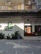 The image depicts the entrance of a building, possibly a mosque, with a sign reading 'Masjid Pogung Dalangan.' There are green plants in pots lining the front, and a few pairs of shoes are visible on the tiled ground, suggesting a place of worship. The upper level features barred windows and a corrugated roof.