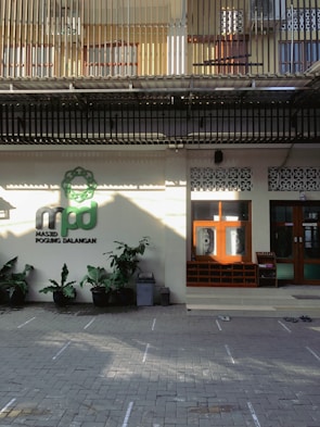 The image depicts the entrance of a building, possibly a mosque, with a sign reading 'Masjid Pogung Dalangan.' There are green plants in pots lining the front, and a few pairs of shoes are visible on the tiled ground, suggesting a place of worship. The upper level features barred windows and a corrugated roof.