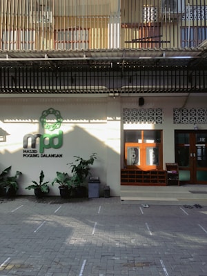 The image depicts the entrance of a building, possibly a mosque, with a sign reading 'Masjid Pogung Dalangan.' There are green plants in pots lining the front, and a few pairs of shoes are visible on the tiled ground, suggesting a place of worship. The upper level features barred windows and a corrugated roof.