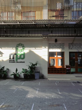 The image depicts the entrance of a building, possibly a mosque, with a sign reading 'Masjid Pogung Dalangan.' There are green plants in pots lining the front, and a few pairs of shoes are visible on the tiled ground, suggesting a place of worship. The upper level features barred windows and a corrugated roof.