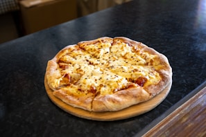 A freshly baked cheese pizza sits on a wooden serving board. The crust is golden brown and thick, with a generous layer of melted cheese sprinkled with chili flakes, hinting at a spicy flavor.