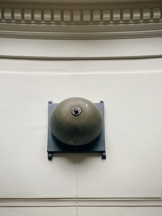 A large metallic bell is mounted on a textured surface with a square base. The background shows intricate architectural molding with a neutral color palette. The bell appears to be aged with a rounded shape and a mechanism at its center.