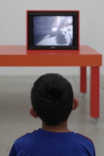 A child is facing away from the camera, watching a television on an orange table in a minimalist, modern setting. The child is wearing a blue shirt, and the television screen shows a blurred image.