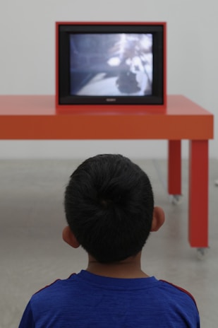 A child is facing away from the camera, watching a television on an orange table in a minimalist, modern setting. The child is wearing a blue shirt, and the television screen shows a blurred image.