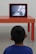 A child is facing away from the camera, watching a television on an orange table in a minimalist, modern setting. The child is wearing a blue shirt, and the television screen shows a blurred image.