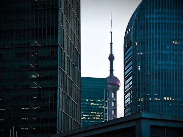 A cityscape featuring tall modern skyscrapers with the distinctive Oriental Pearl Tower prominently positioned between them. The scene is captured during a time when lighting within the buildings enhances the structure contrasts. The sky appears dim, suggesting twilight or early evening, adding a serene ambiance.