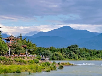 A scenic landscape features a traditional Asian-style pagoda surrounded by lush greenery. In the background, rolling blue mountains under a partially cloudy sky provide a serene backdrop. A river flows gently in the foreground, adding to the tranquil setting.