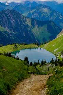 A scenic mountain landscape with a clear, blue lake surrounded by lush greenery and pine trees. The rugged mountains in the background rise up against a sky with scattered clouds. A dirt path leads the viewer's eye towards the lake, creating a sense of depth and adventure.