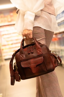 A person is holding a burgundy leather handbag with a textured finish. The hand is adorned with a large ring, and the individual is dressed in plaid pants and a sheer white blouse with voluminous sleeves. The background appears to be a grocery store aisle with shelves and blurred products.