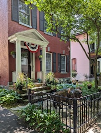 A charming red-brick townhouse features a small front porch adorned with a decorative flag. Potted plants line the steps, and a neatly landscaped garden includes hostas and other greenery. A black metal fence encloses a sculpture resembling a buffalo. The scene is shaded by a large tree with bright green leaves, casting dappled shadows.