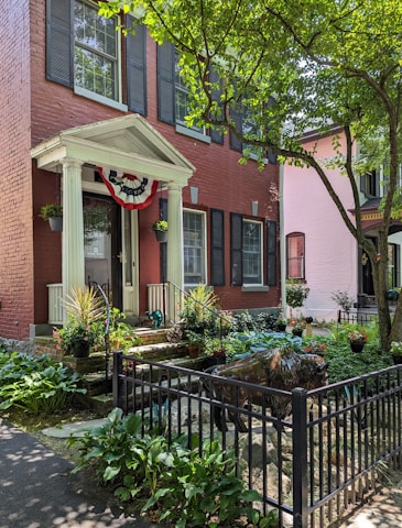 A charming red-brick townhouse features a small front porch adorned with a decorative flag. Potted plants line the steps, and a neatly landscaped garden includes hostas and other greenery. A black metal fence encloses a sculpture resembling a buffalo. The scene is shaded by a large tree with bright green leaves, casting dappled shadows.