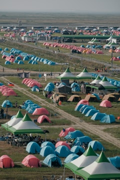 A large number of brightly colored tents are set up in a sprawling outdoor area, arranged in rows on green grass. There are paths between the tents and some canopy-covered seating areas with people gathered underneath. The area appears to be a campsite or festival setting with various colorful tents, possibly indicating a large event or gathering.