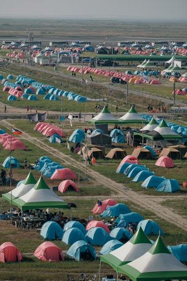 A large number of brightly colored tents are set up in a sprawling outdoor area, arranged in rows on green grass. There are paths between the tents and some canopy-covered seating areas with people gathered underneath. The area appears to be a campsite or festival setting with various colorful tents, possibly indicating a large event or gathering.