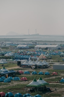 A large outdoor camping area with numerous tents arranged in rows on a grassy field. White canopy tents are also set up, likely for communal gatherings or events. In the background, there are two wind turbines and a hazy sky overlooking a distant shoreline.