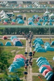 Rows of vibrant blue and pink tents are set up in a large open area, likely for an outdoor event or gathering. People are walking along dirt paths between the tents, holding black umbrellas. There are large canopies in the background with a sign that reads 'Scout', and buses are parked further back.