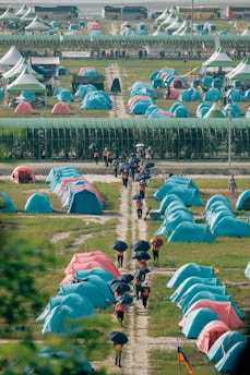 Rows of vibrant blue and pink tents are set up in a large open area, likely for an outdoor event or gathering. People are walking along dirt paths between the tents, holding black umbrellas. There are large canopies in the background with a sign that reads 'Scout', and buses are parked further back.