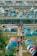 Rows of vibrant blue and pink tents are set up in a large open area, likely for an outdoor event or gathering. People are walking along dirt paths between the tents, holding black umbrellas. There are large canopies in the background with a sign that reads 'Scout', and buses are parked further back.