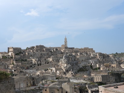 A historical townscape built into a rugged hill with numerous stone buildings, featuring an ancient church tower in the center against a bright blue sky. The architecture reflects a harmonious blend with the natural limestone rock formations.