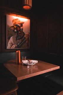 A dimly lit dining booth with a wooden table and two empty glasses. A rolled napkin lies on a small plate, and a menu stands upright on the table. Above the booth, a piece of art features a person with colorful spheres obscuring their face, contributing to an intimate and mysterious atmosphere.