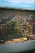 A view of a suburban house seen through a window. The house has a modern architectural design with a gable roof and large front porch. There are trees, a well-maintained lawn, and a black sign in the front yard. Two garbage bins are placed near the sidewalk, with a parked car visible to the right.