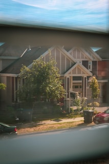 A view of a suburban house seen through a window. The house has a modern architectural design with a gable roof and large front porch. There are trees, a well-maintained lawn, and a black sign in the front yard. Two garbage bins are placed near the sidewalk, with a parked car visible to the right.