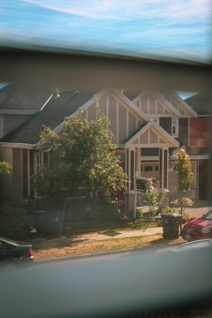 A view of a suburban house seen through a window. The house has a modern architectural design with a gable roof and large front porch. There are trees, a well-maintained lawn, and a black sign in the front yard. Two garbage bins are placed near the sidewalk, with a parked car visible to the right.