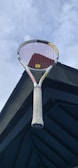 A tennis racket with a white handle and red strings is positioned against a dark architectural structure and a cloudy sky.
