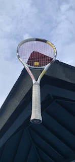 A tennis racket with a white handle and red strings is positioned against a dark architectural structure and a cloudy sky.