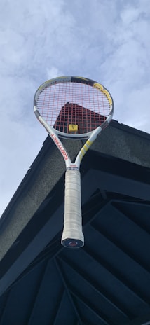 A tennis racket with a white handle and red strings is positioned against a dark architectural structure and a cloudy sky.