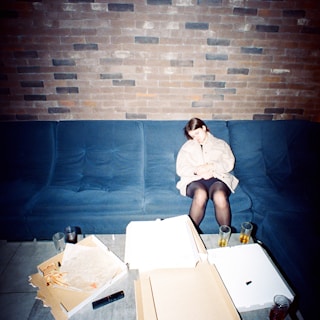 A person is sitting on a blue couch with their eyes closed, leaning slightly forward. The setting features a brick wall in the background. On the table in front of them, there are empty pizza boxes, a remote control, and several glasses with drinks, indicating a recent meal or gathering.
