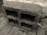 A concrete cinder block with rough texture positioned on a paved surface. The block has three large rectangular openings and is placed against a stone or concrete wall in an outdoor setting.