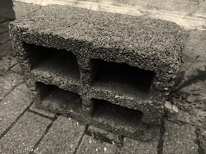 A concrete cinder block with rough texture positioned on a paved surface. The block has three large rectangular openings and is placed against a stone or concrete wall in an outdoor setting.