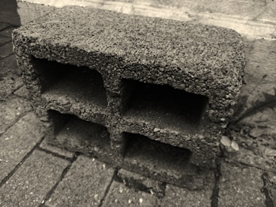 A concrete cinder block with rough texture positioned on a paved surface. The block has three large rectangular openings and is placed against a stone or concrete wall in an outdoor setting.