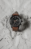 A wristwatch with a brown leather strap and a black, detailed dial face. It features multiple dials and numerals, displayed on a textured, light-colored surface.
