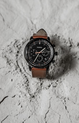 A wristwatch with a brown leather strap and a black, detailed dial face. It features multiple dials and numerals, displayed on a textured, light-colored surface.