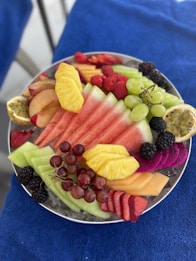 A colorful assortment of fresh fruits is arranged on a plate. The selection includes watermelon, pineapple slices, grapes, blackberries, raspberries, strawberries, cantaloupe, passion fruit halves, dragon fruit slices, and apple slices. The fruits are neatly displayed on a bed of ice, creating a refreshing and appetizing presentation, with the plate placed on a blue textured surface.