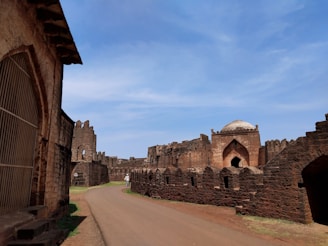 An ancient stone fortress with high walls and a central dome structure in the middle. The path leading up to the fortress is surrounded by green grass, and the sky above is clear with a few clouds. The walls are made of red-brown stone and show signs of age and wear.