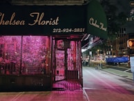 A florist shop named 'Chelsea Florist' with a dark green awning and a neon pink light illuminating the interior visible through the shop's window. The shop appears to be closed, with metal grates pulled over the entrance and window. The background shows a dimly lit city street at night, including a sidewalk, street signs, and various urban buildings.